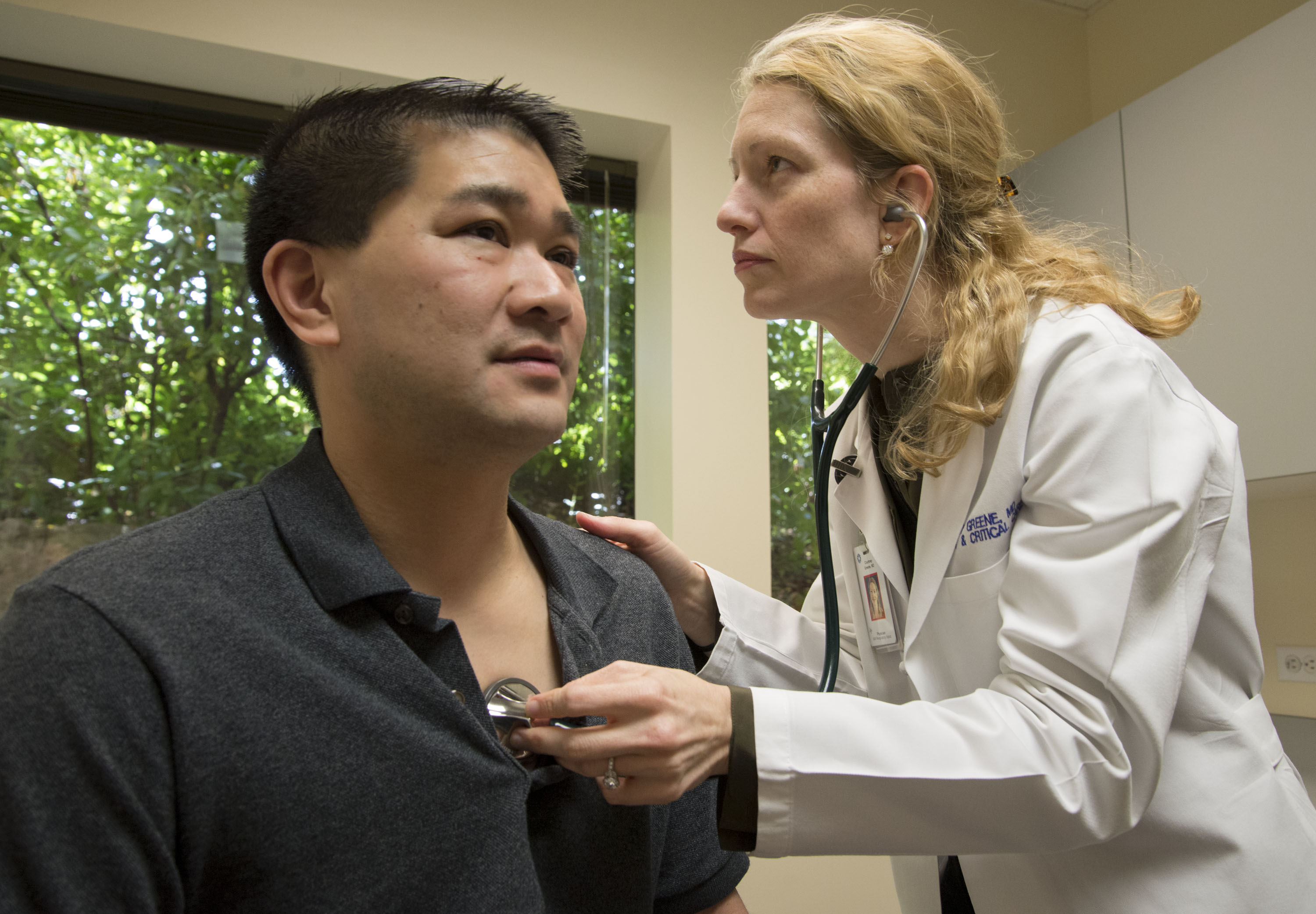 Male patient with black hair being examined with a stethoscope by a female physician with blonde hair