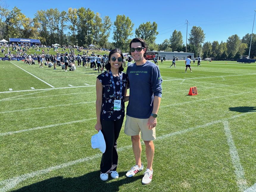 Two people smile at the camera, with a football practice behind them.