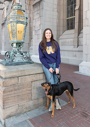 A woman in a purple shirt with a gold W holds the leash of a medium-size black and tan dog.