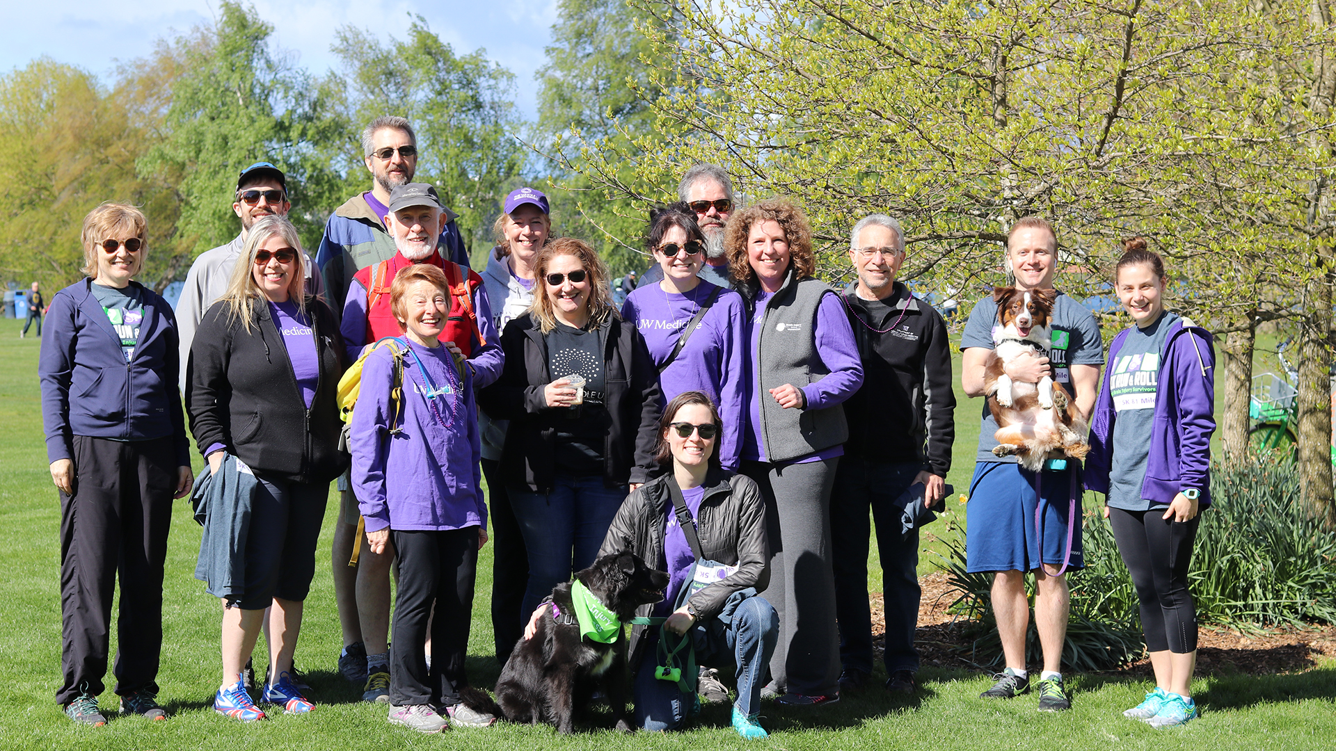 15 people in purple t-shirts (and two dogs) stand outside on a sunny fall day