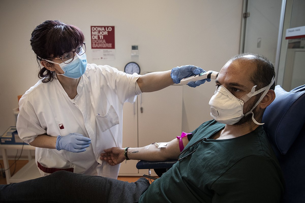 Male masked COVID-19 patient in exam chair getting forehead temperature measured by a masked nurse in white lab coat