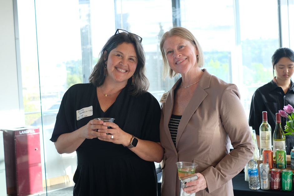 Two women smile at the camera in front of a large window.