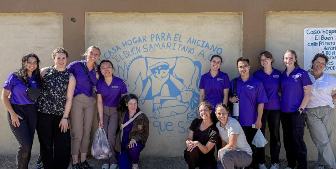 A group of people, most in purple shirts, pose again a wall with a mural of the Good Samaritan and writing in Spanish.