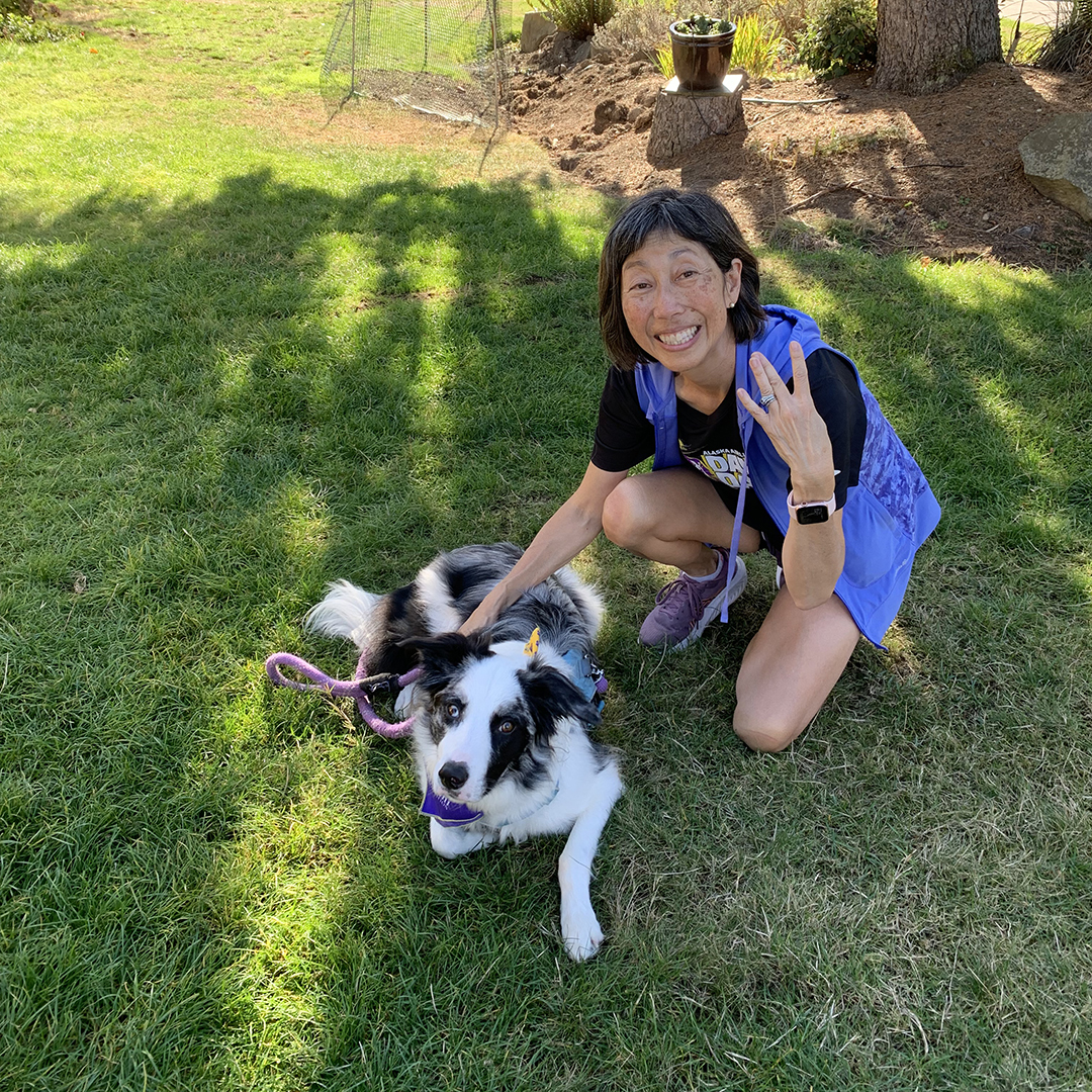 A woman and a black and white dog kneel in a grassy yard.