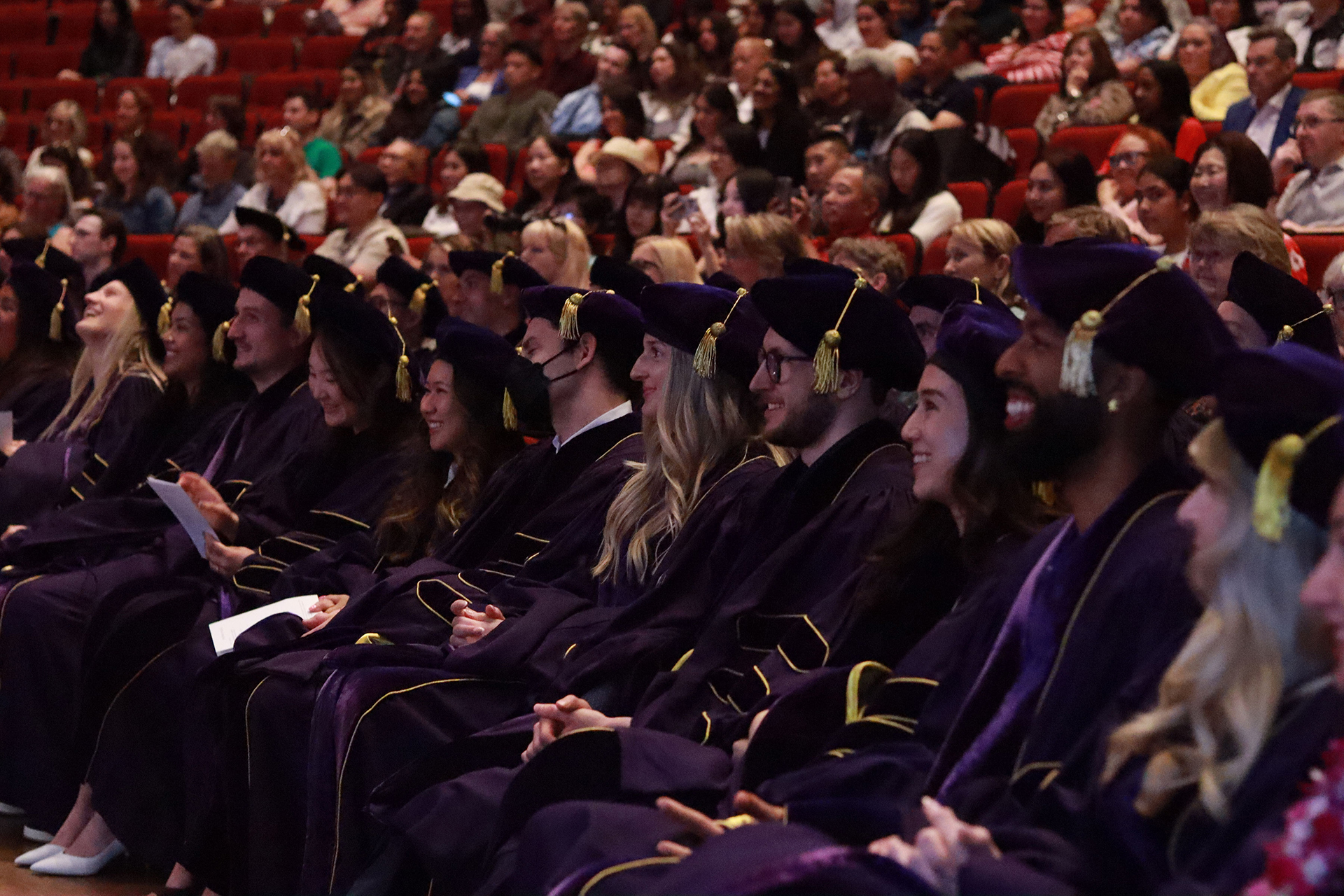 A group of graduates in gowns sit in a full auditorium.
