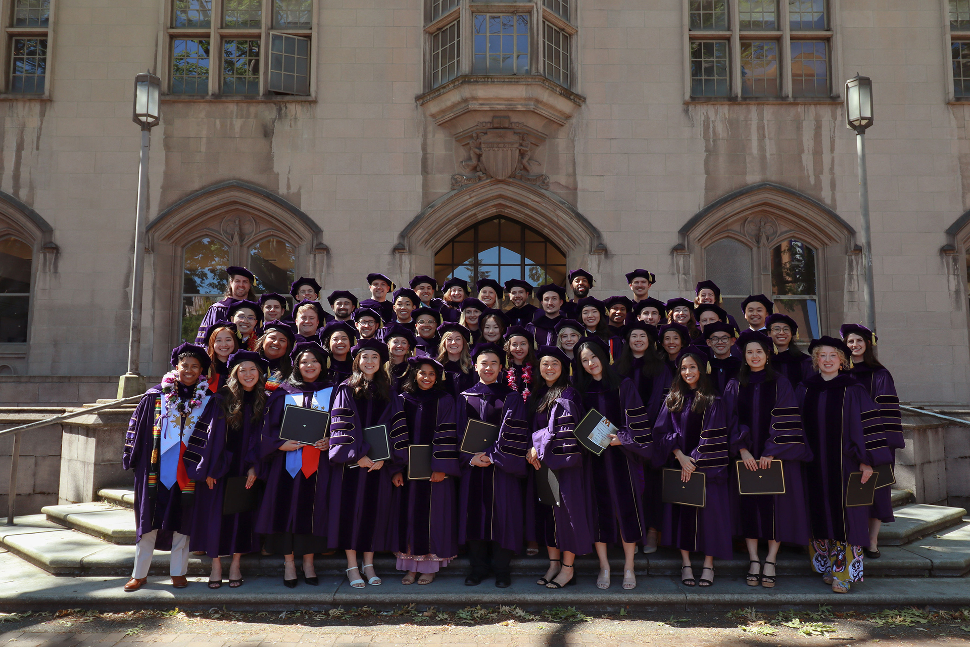 A large group of graduates in gowns pose in front of a stone building.