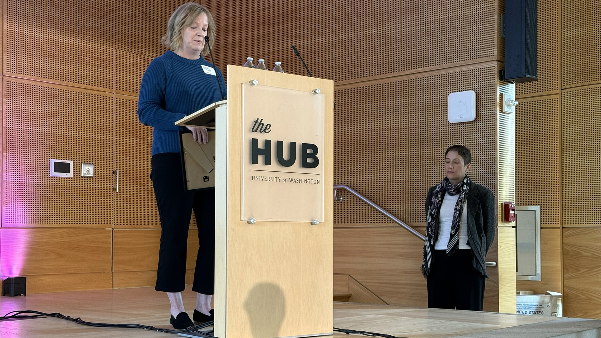 A woman speaks at a podium, while another woman waits to come on stage.