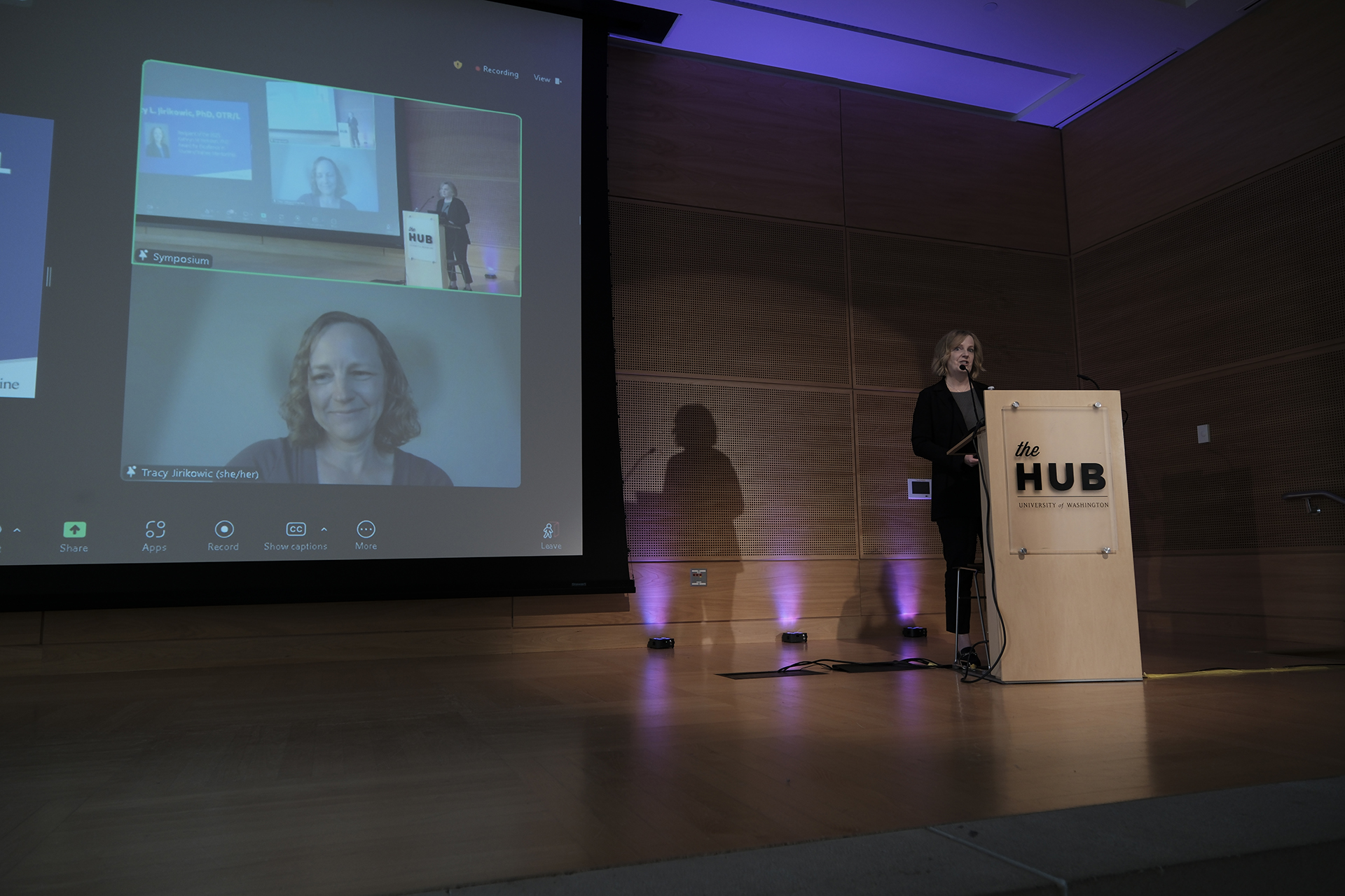 Dawn Edhe stands on stage, while a projector screen shows Tracy Jirikowic on Zoom.