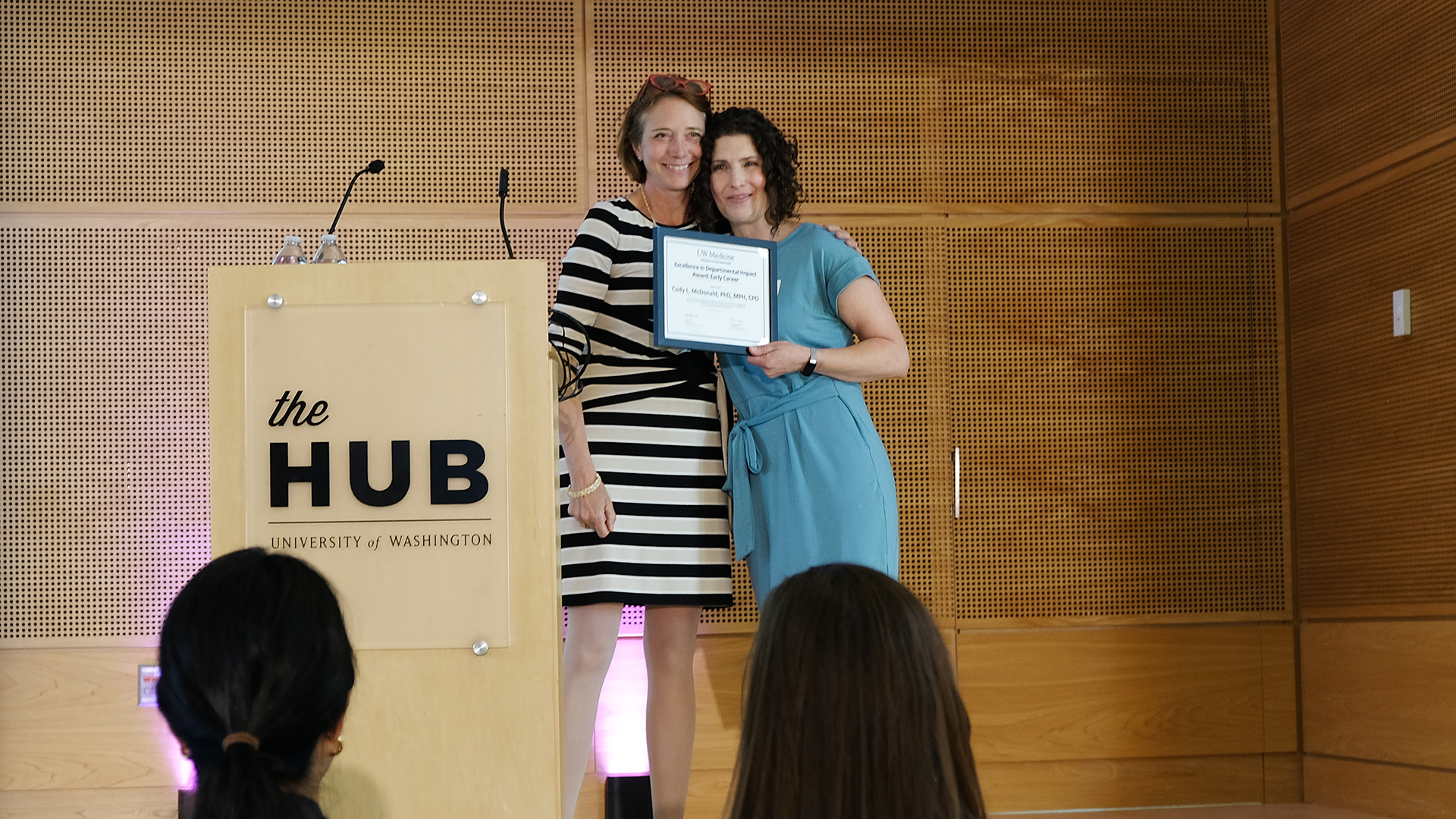 Two women smile on stage while one holds a certificate