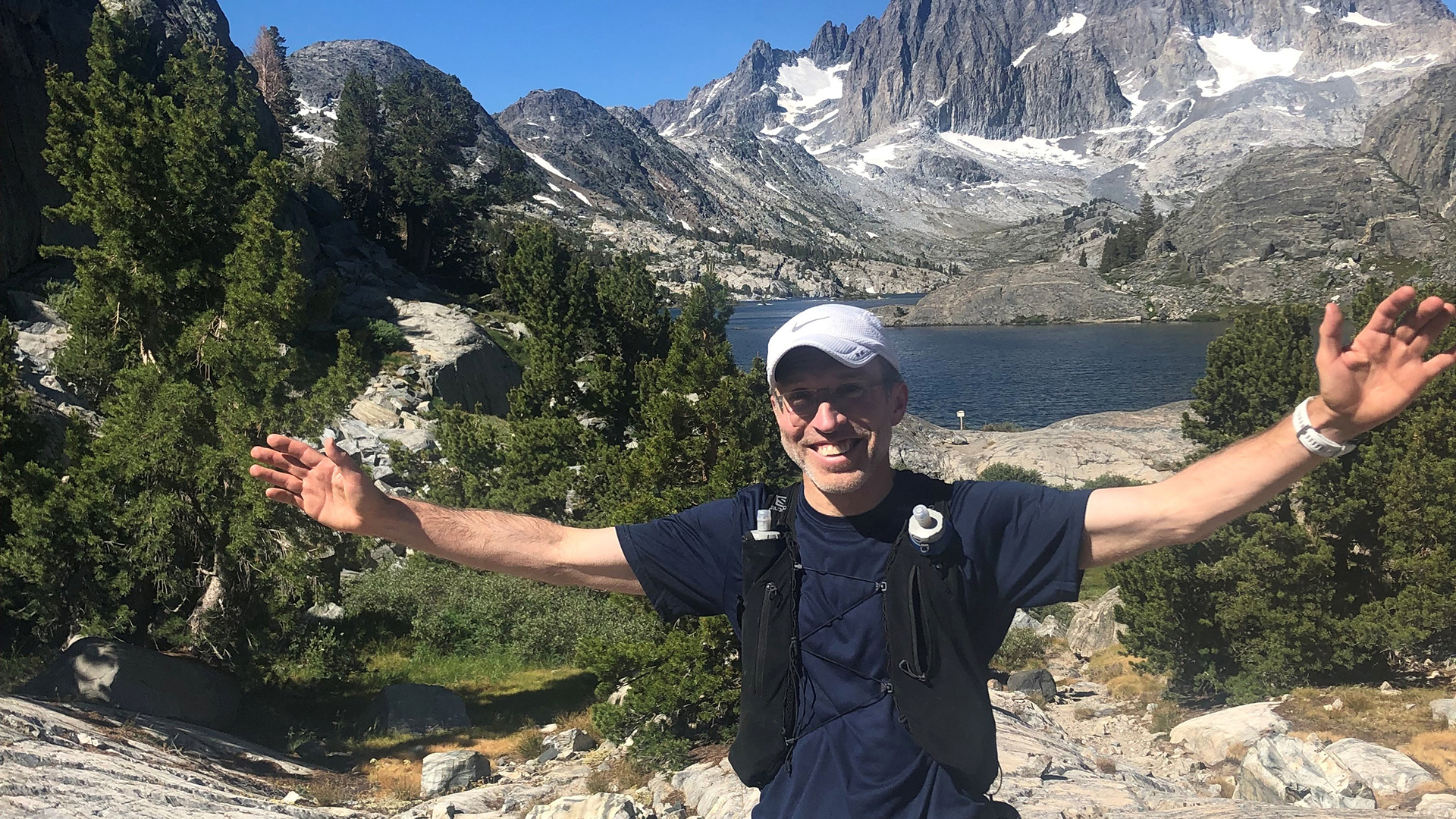 Dr. Harrast spreads his arms to celebrate the gorgeous mountain and lake behind him.