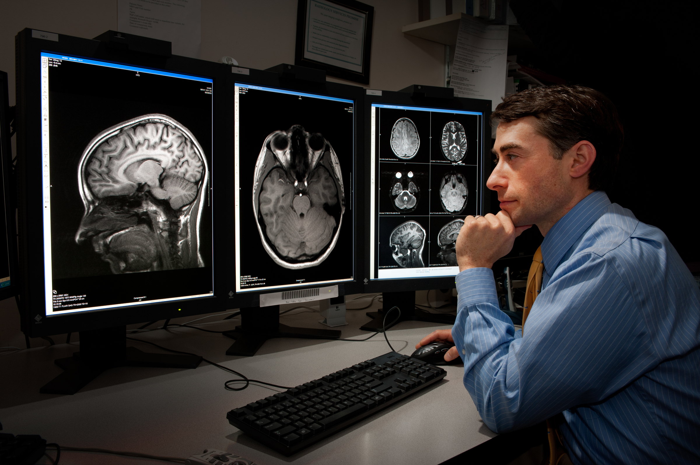 Male scientist in blue dress shirt sitting at a desk looking at three monitors with images of brain scans