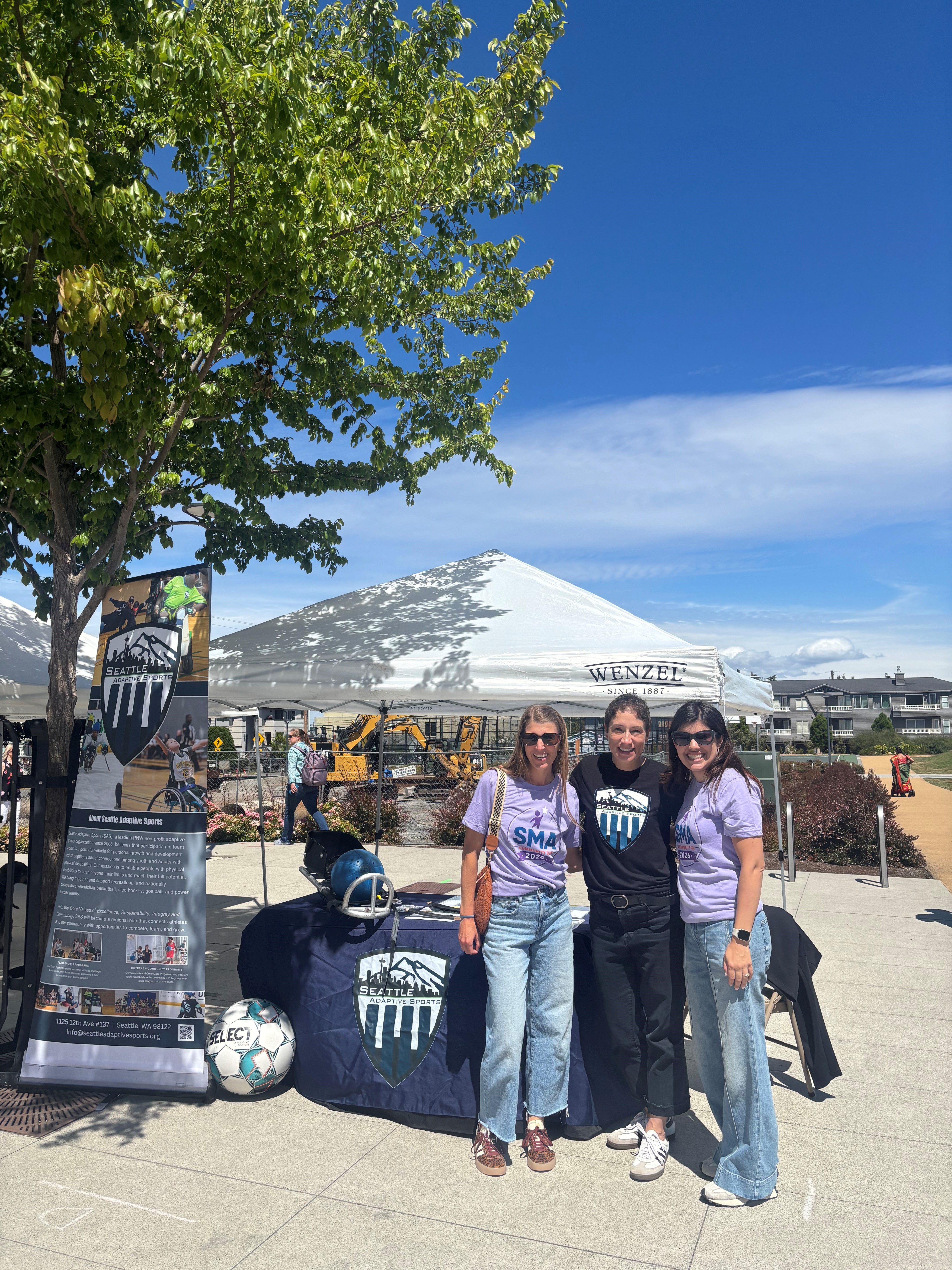 Three women stand in front of a Seattle Adaptive Sports display at an outdoor event on a sunny day.
