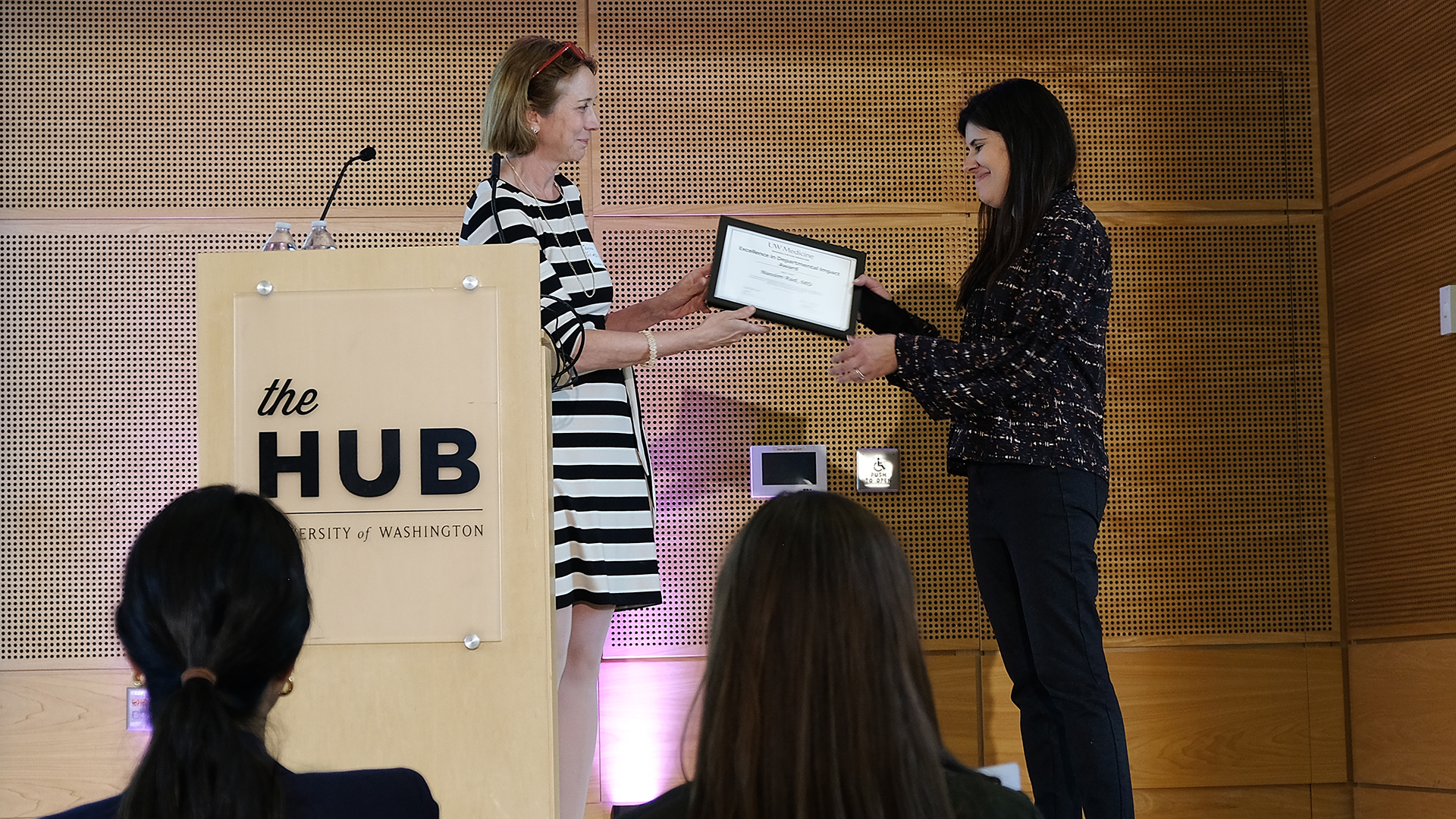 A woman hands a certificate to another woman on an event stage.
