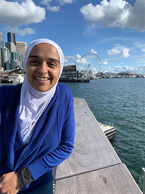 Rehab Abuatiq stands on a pier in Seattle on a sunny day, with a ferris wheel in the background.