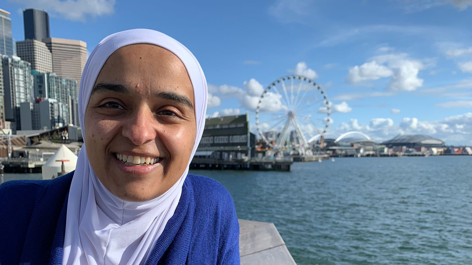 Reham, wearing a hijab and a blue sweater, stands at the Seattle waterfront on a sunny day.