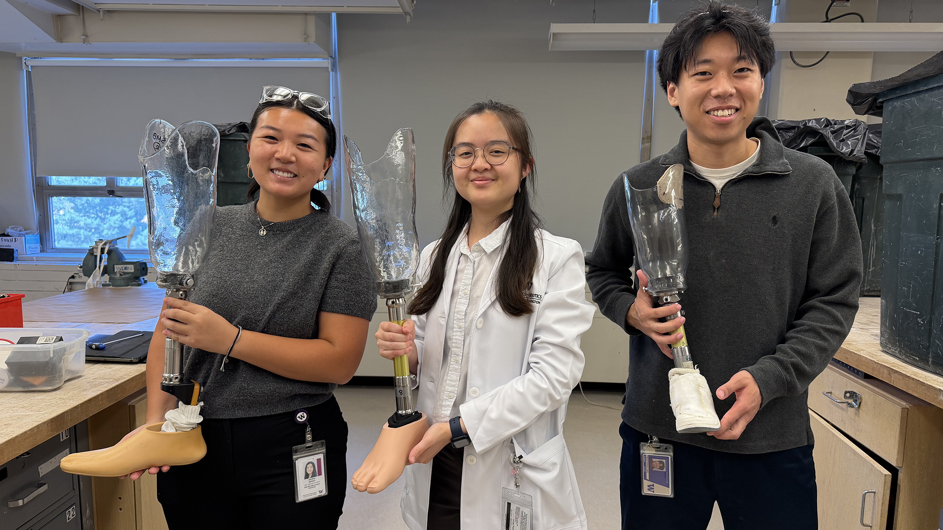 Three students stand in a lab holding prosthetic lower legs.