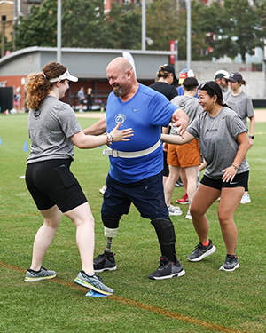 Two women assist an athlete with a lower limb prosthetic on a playing field.