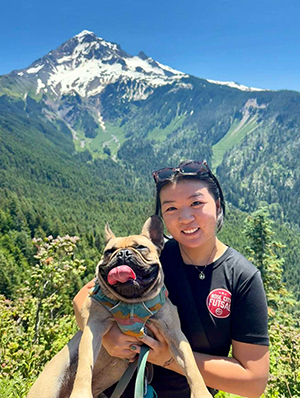 A woman holds a dog outside with a mountain rising behind her.