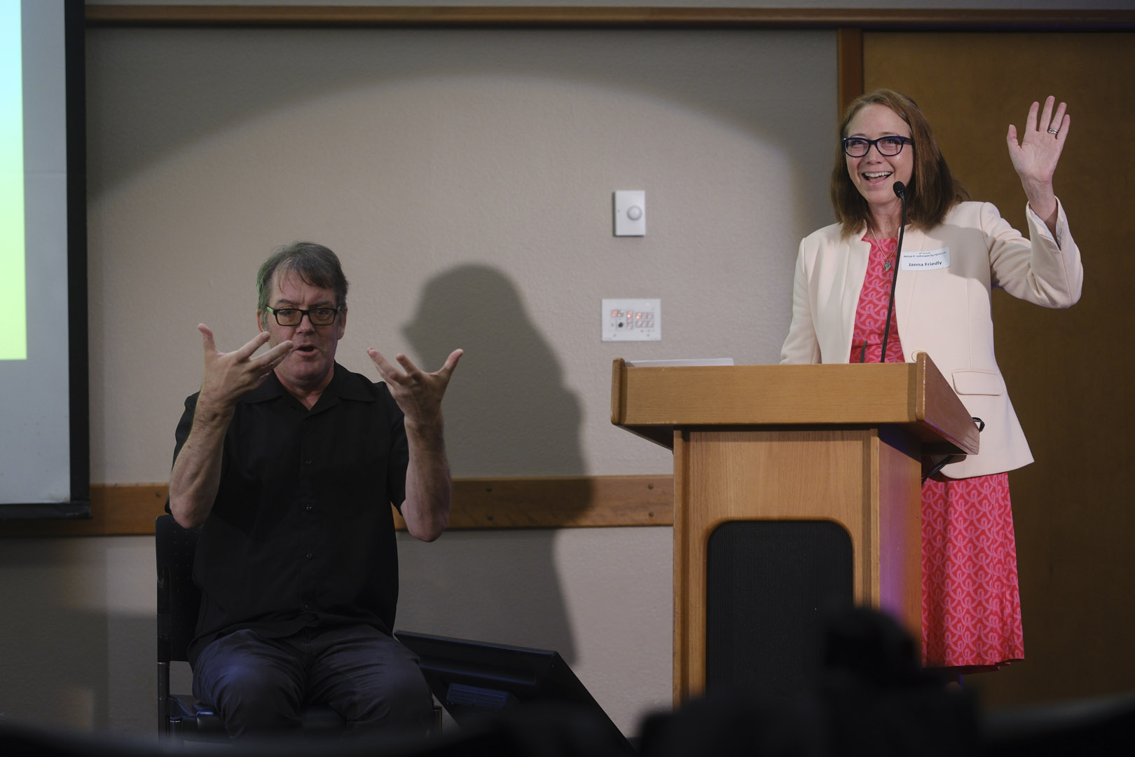 Janna Friedly waves to the crowd. An ASL interpreter sits next to her.