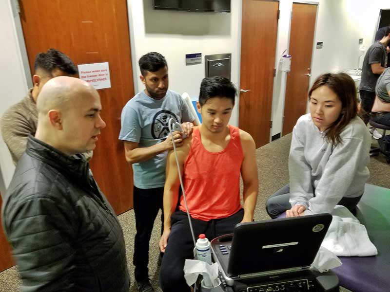 A man uses a device on another's shoulder, while three people look on at a computer screen.