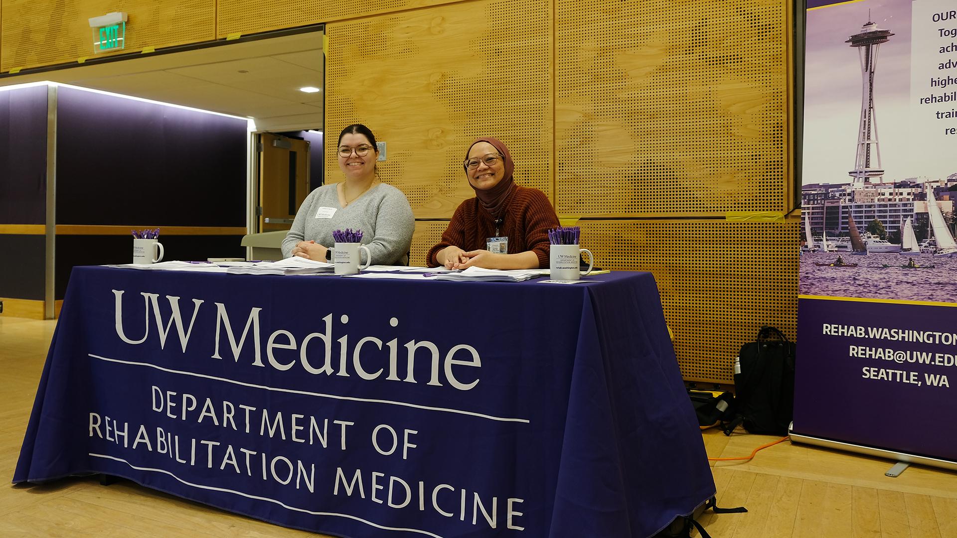 Two women smile at a reception desk covered in a purple tablecloth.