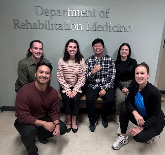 A group of 6 people pose in front of a Department of Rehabilitation Medicine sign.