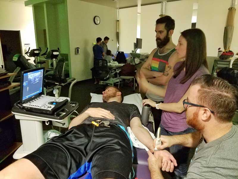 A group watches a monitor as one uses a butterfly probe on another's wrist. 
