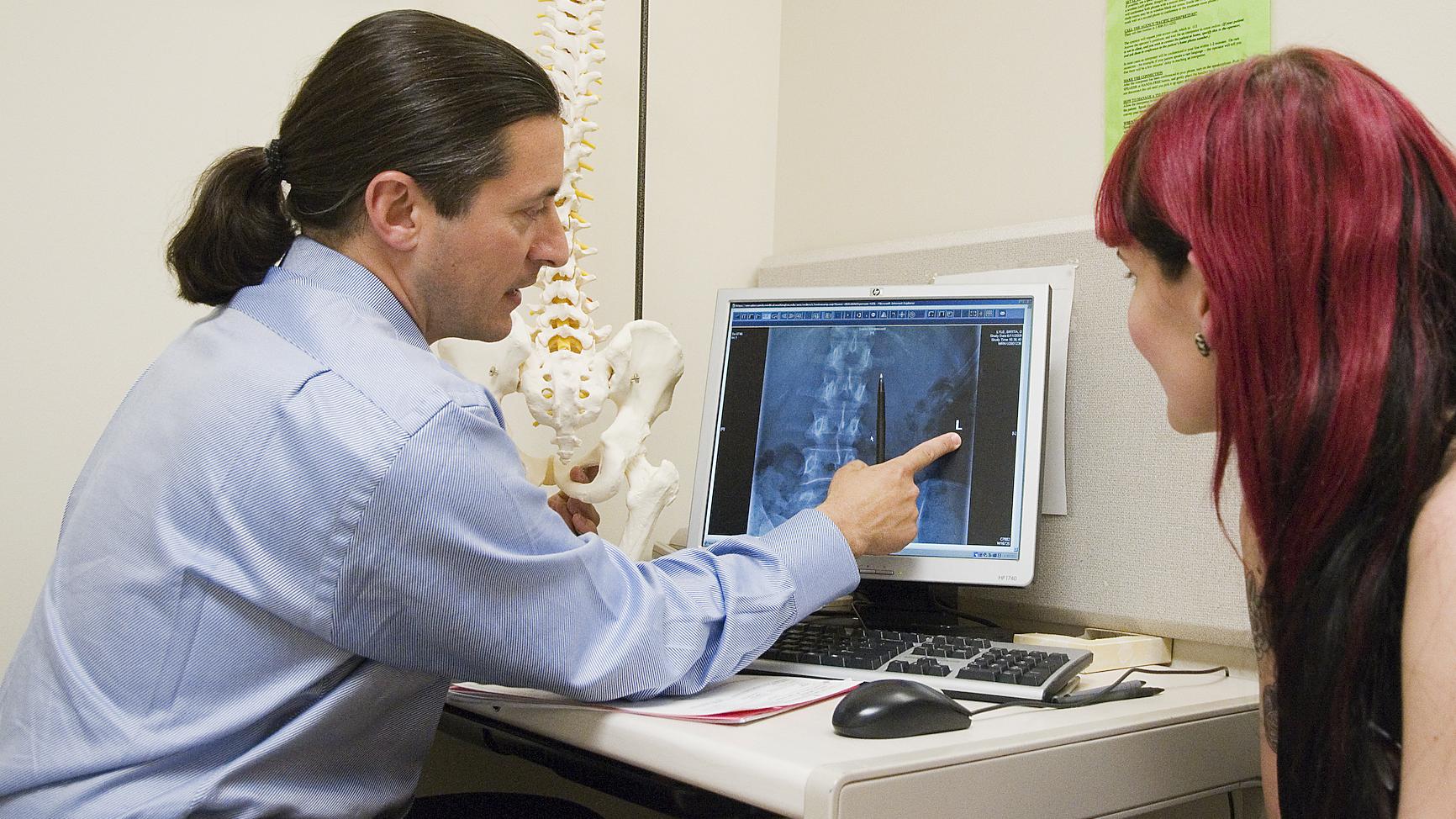 A doctor talks to a patient while pointing at a spinal X-ray.
