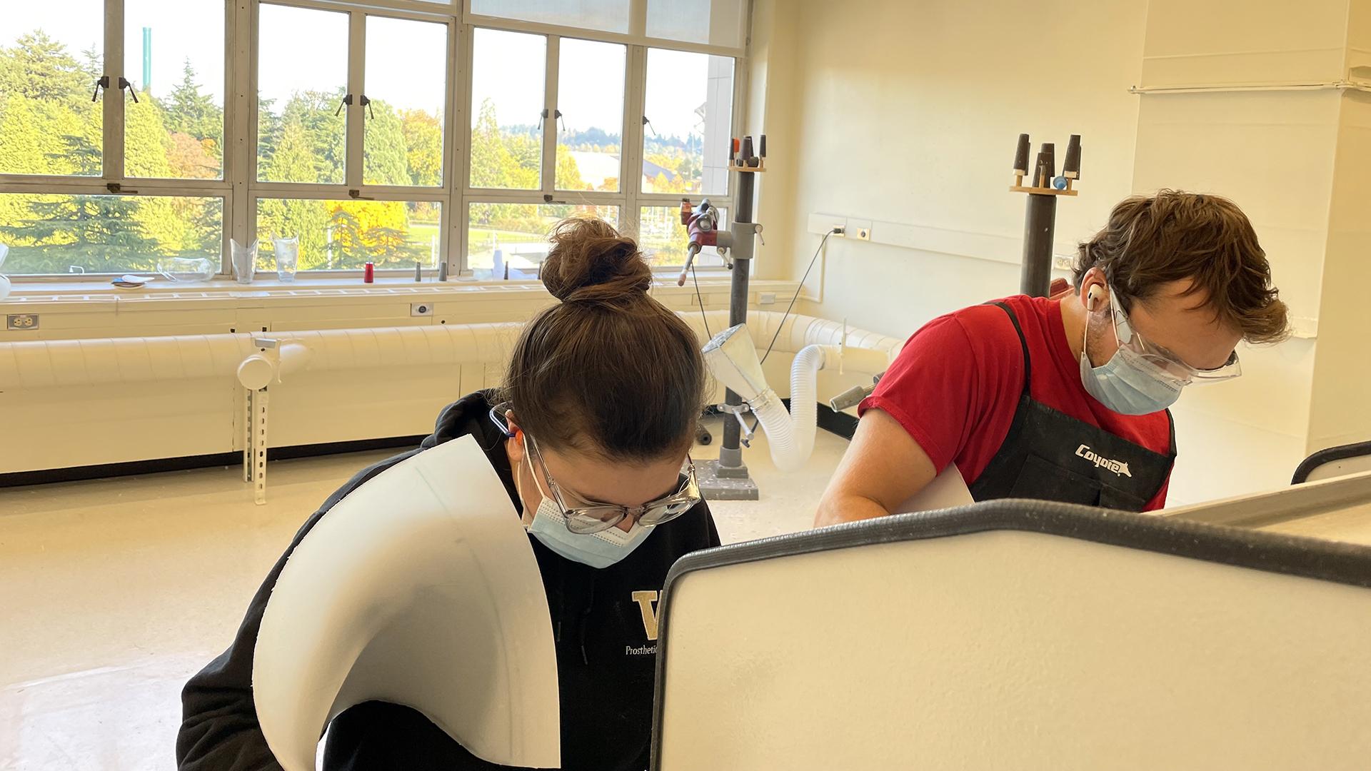 Two students wearing masks bend over a worktable in a sun-lit lab. 