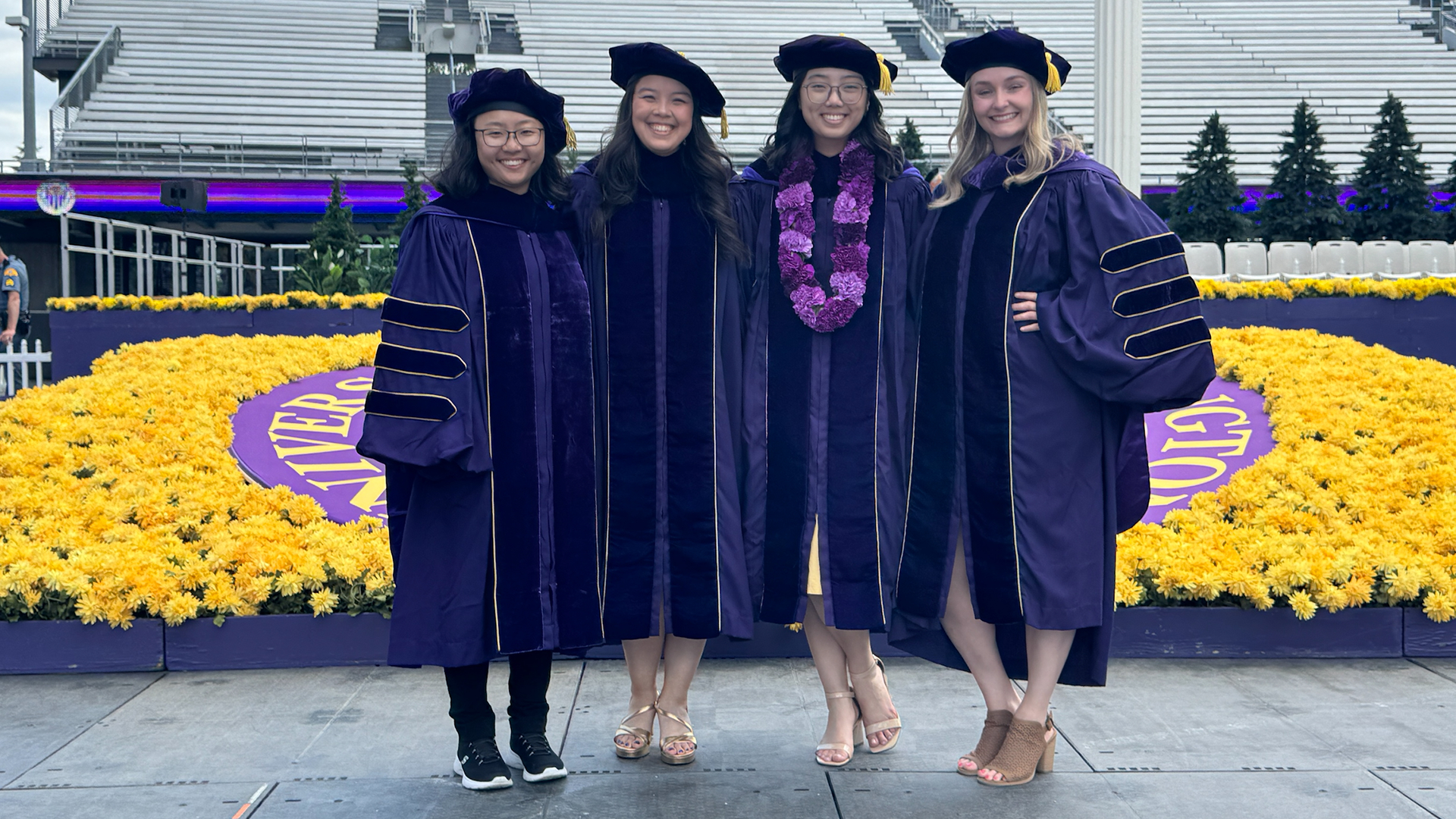 Four women in purple graduation robes stand in front of yellow flowers. 