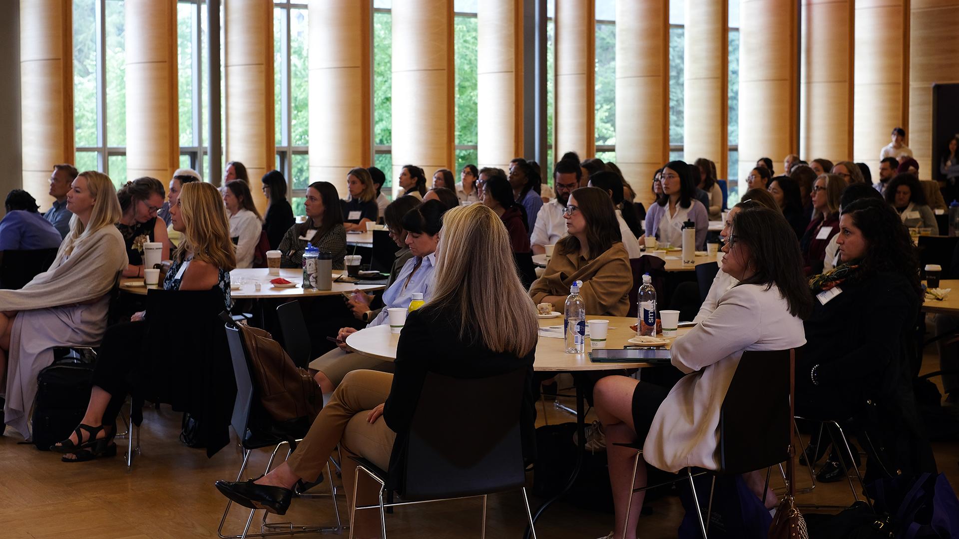 A crowd in fills a conference room, with a bank of windows in the background.