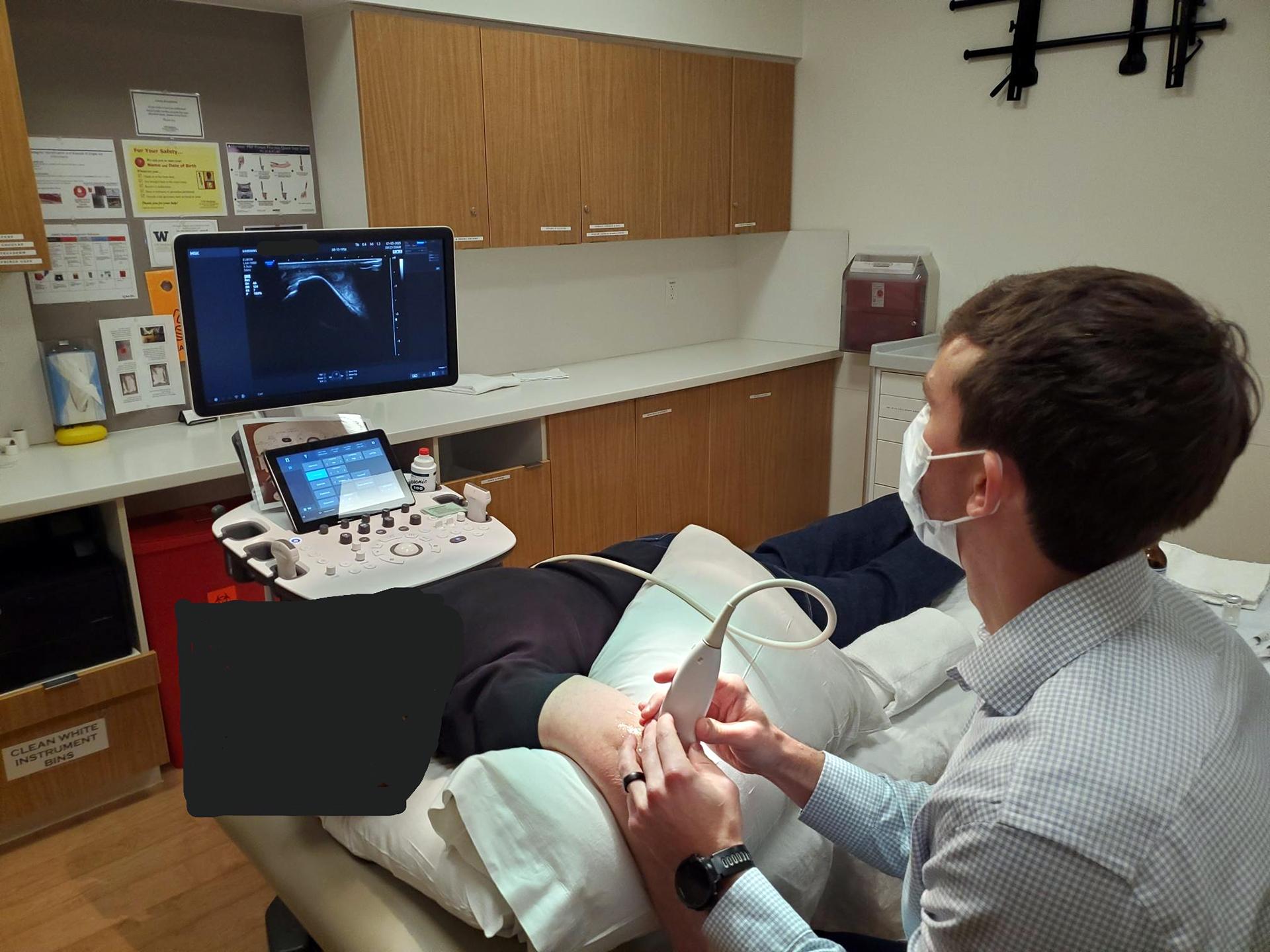 A patient lies on a treatment bed while a doctor checks her arm with an ultrasound probe. 