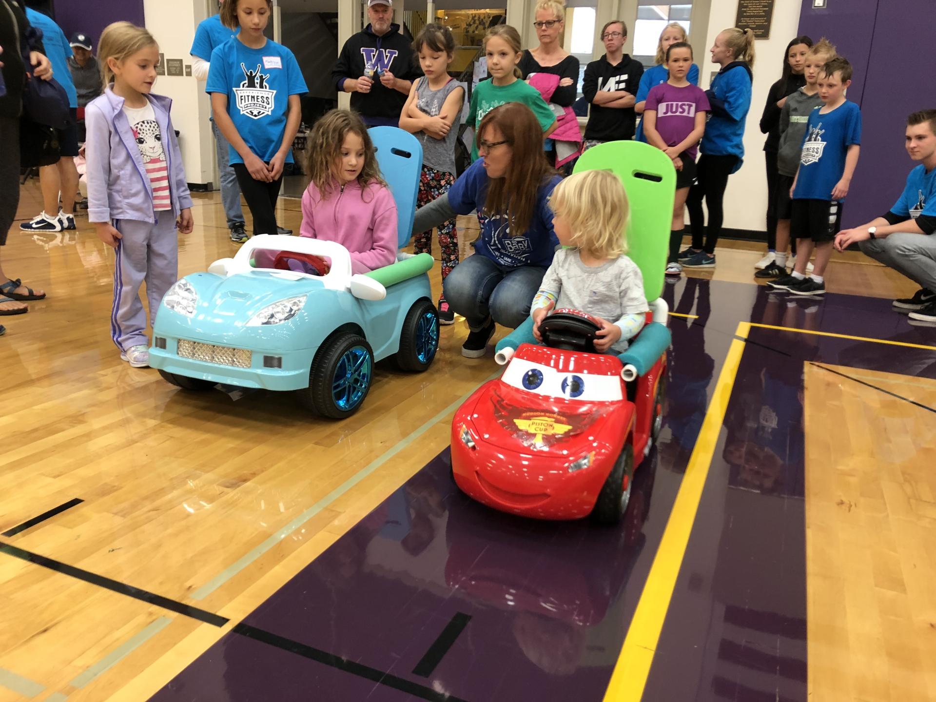 Two Go Baby Go! carts driven by two girls who are surrounded by other children and a female adult