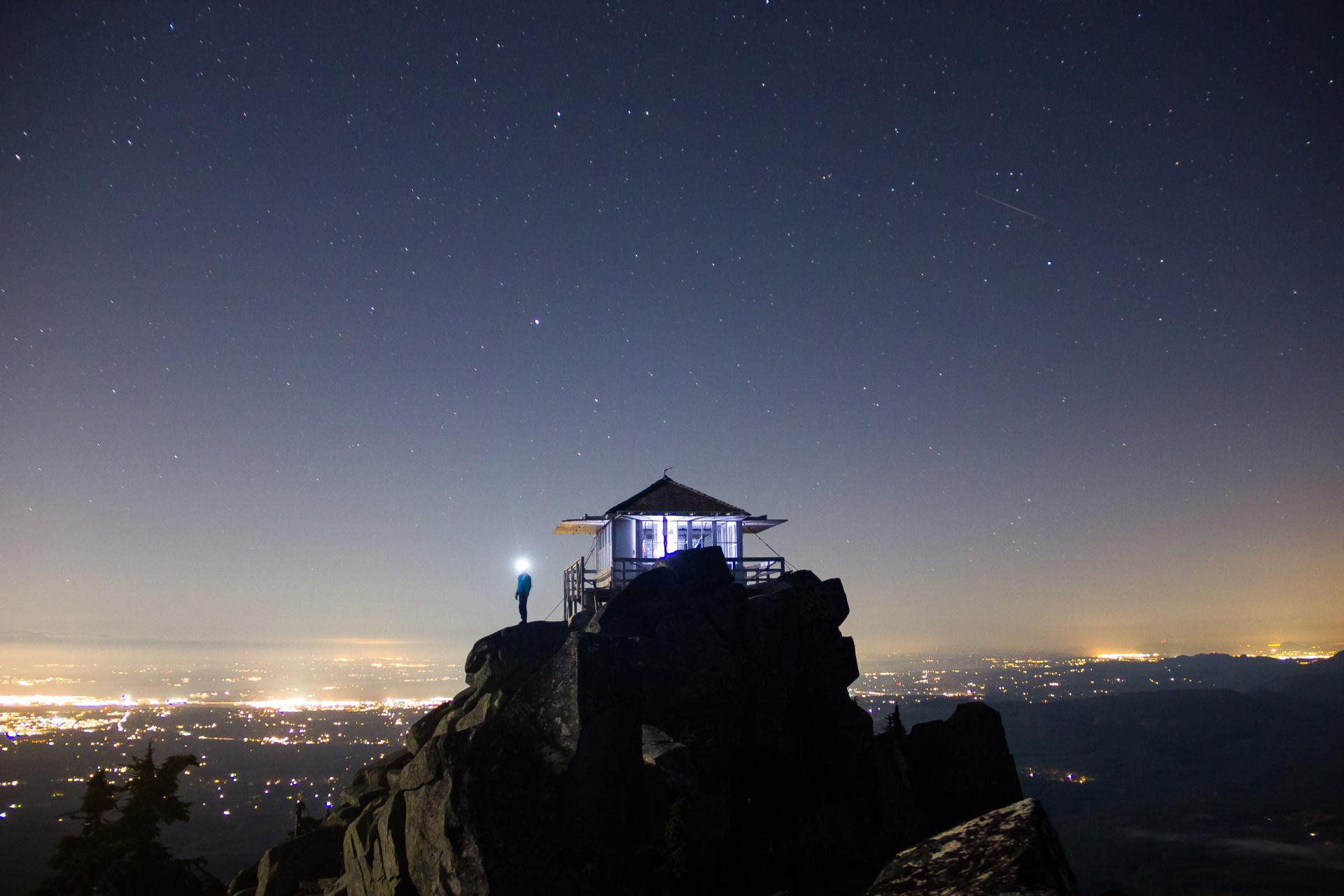 Hiker standing atop mountain lookout at night with lights in the distance