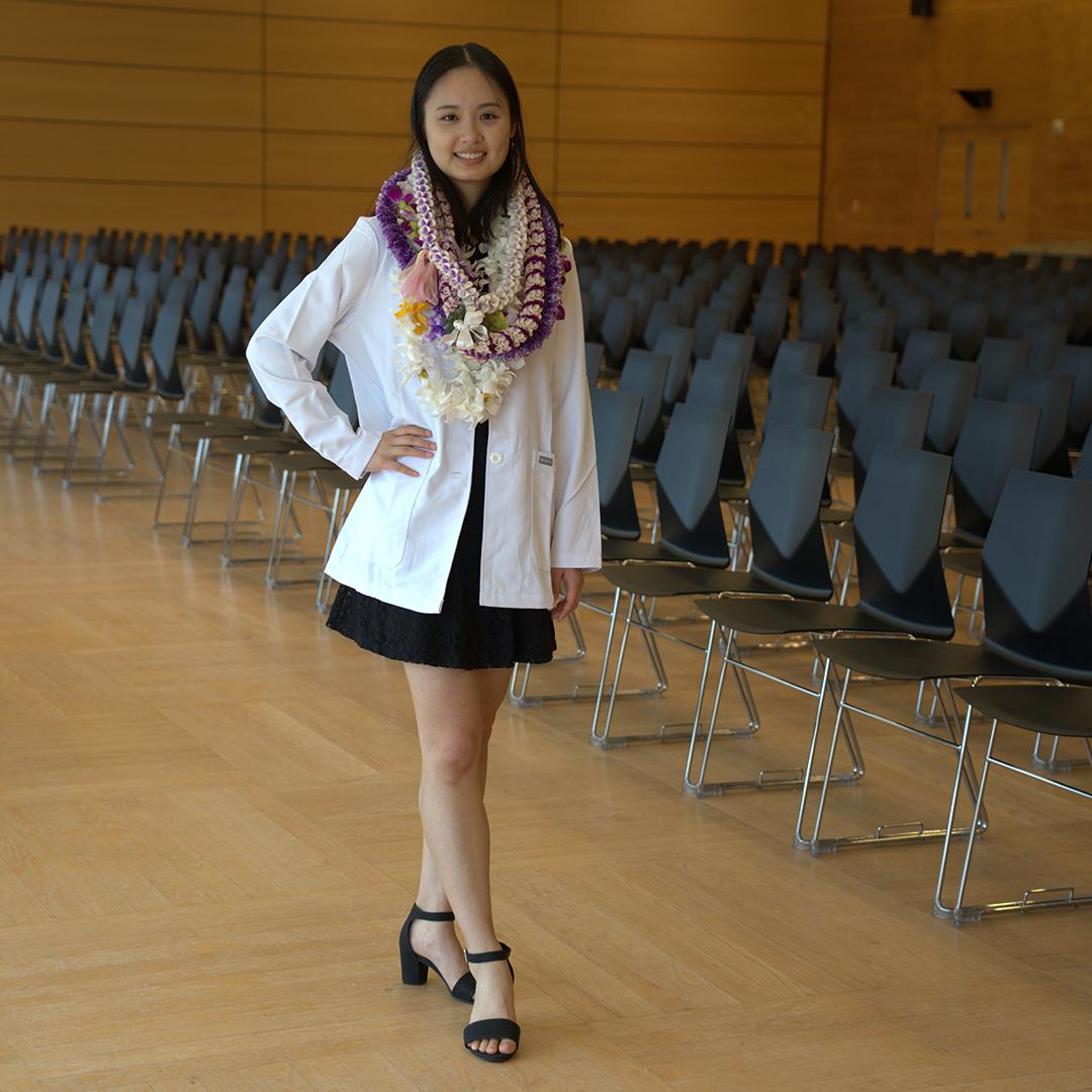 A student poses on stage wearing a white coat and Hawaian leis. 