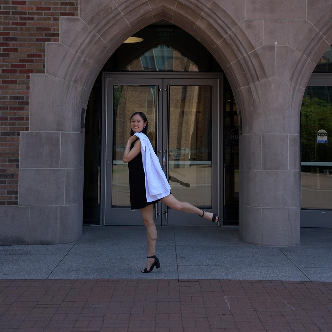 A woman in a black dress with a white coat over her shoulder stands in an attitude derrière next to a brick building. 
