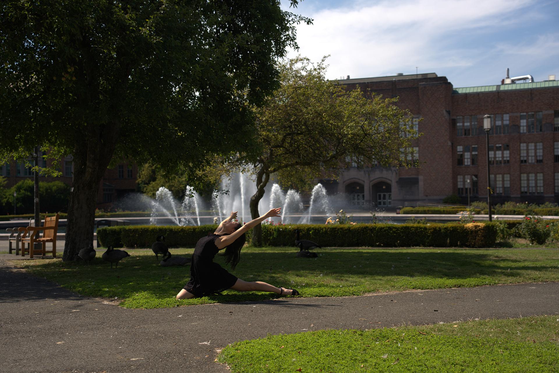 A woman in a black dress lunges and throws up her hands dramatically in front of a fountain. 