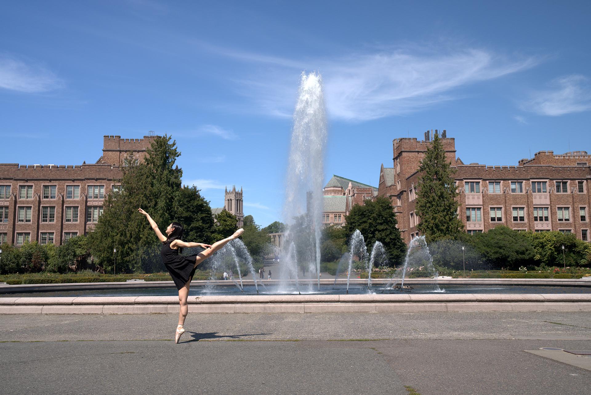 A woman in a black dress does an arabesque in front of a high fountain on a sunny day. 