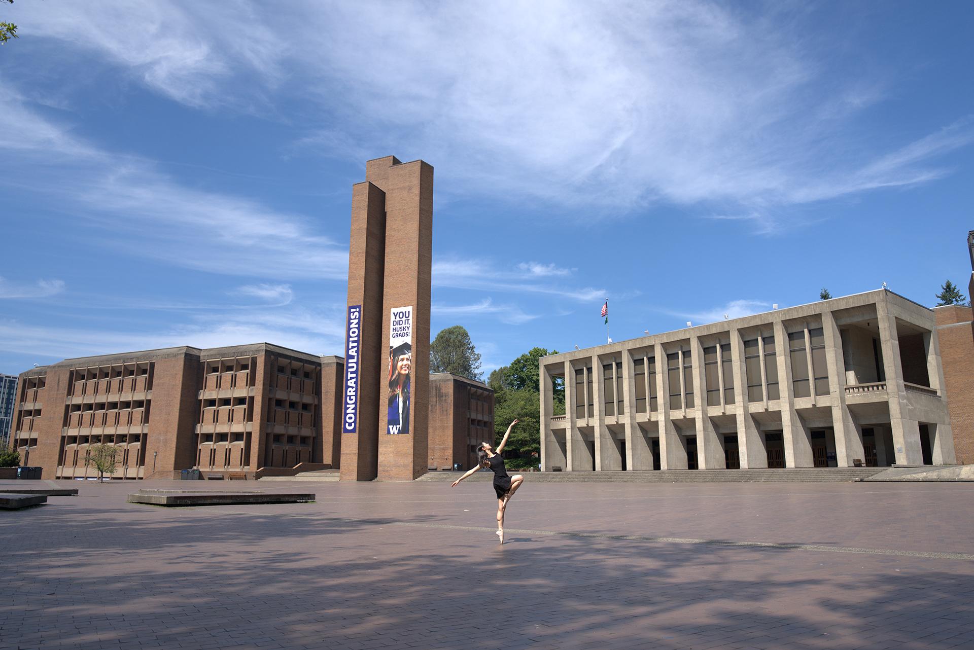 A woman in a black dress dances in a brick square with a brick tower in the background. 