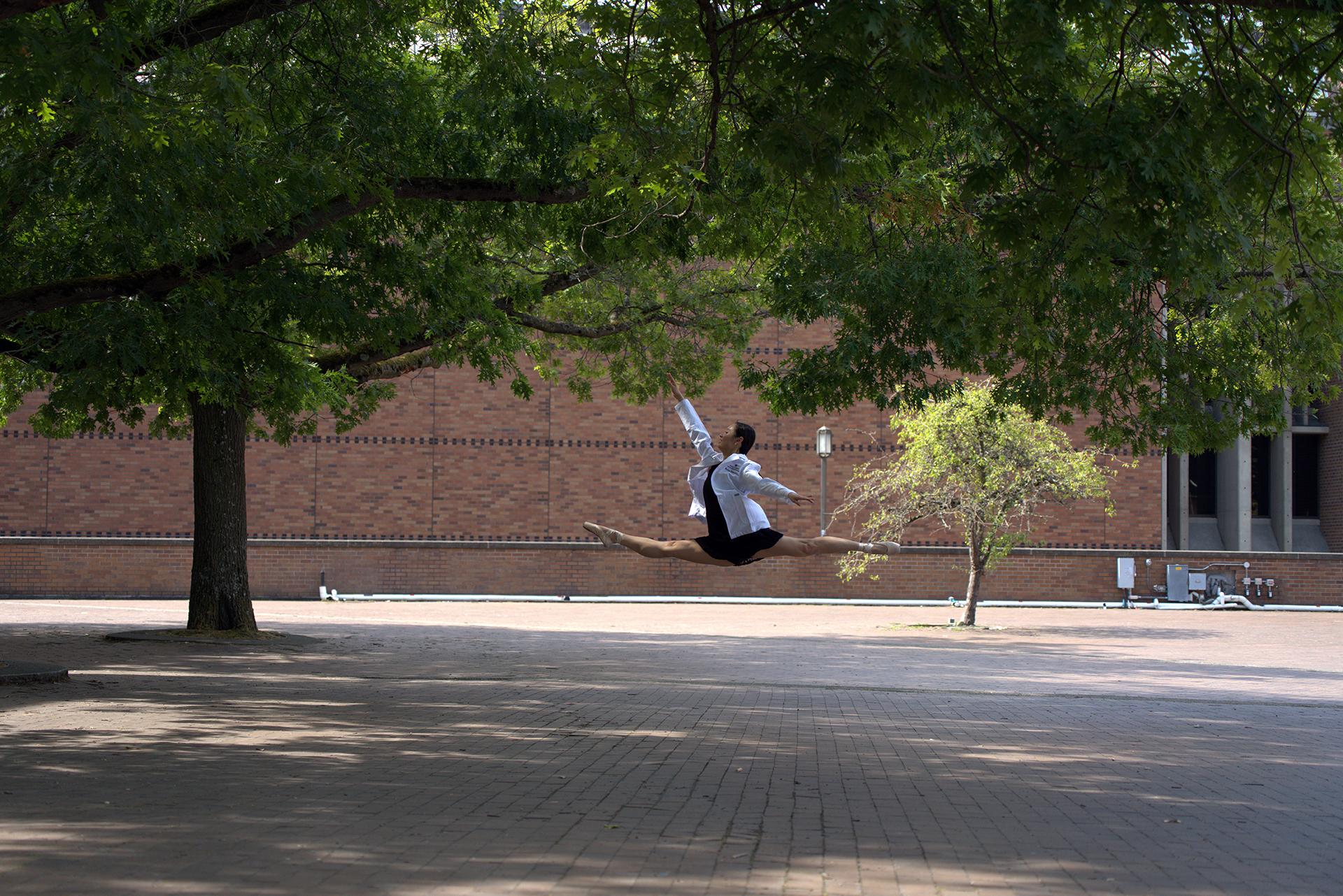 A woman in a black dress and a white coat leaps dramatically in a red brick plaza. 