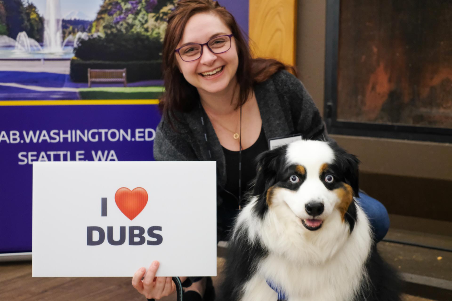 A woman smiles and holds a sign that says, "I heart Dubs" next to a very fluffy dog with big round eyes. 