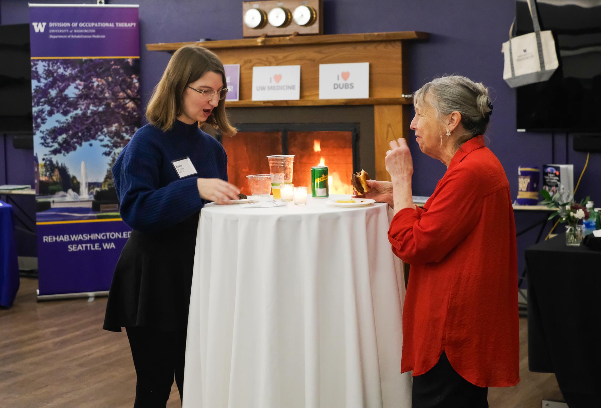 Two women talk over a white-draped high top table. 