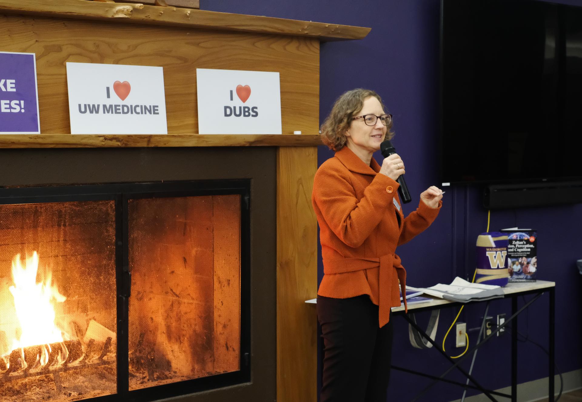 A woman in an orange jacket speaks in front of a fireplace. 