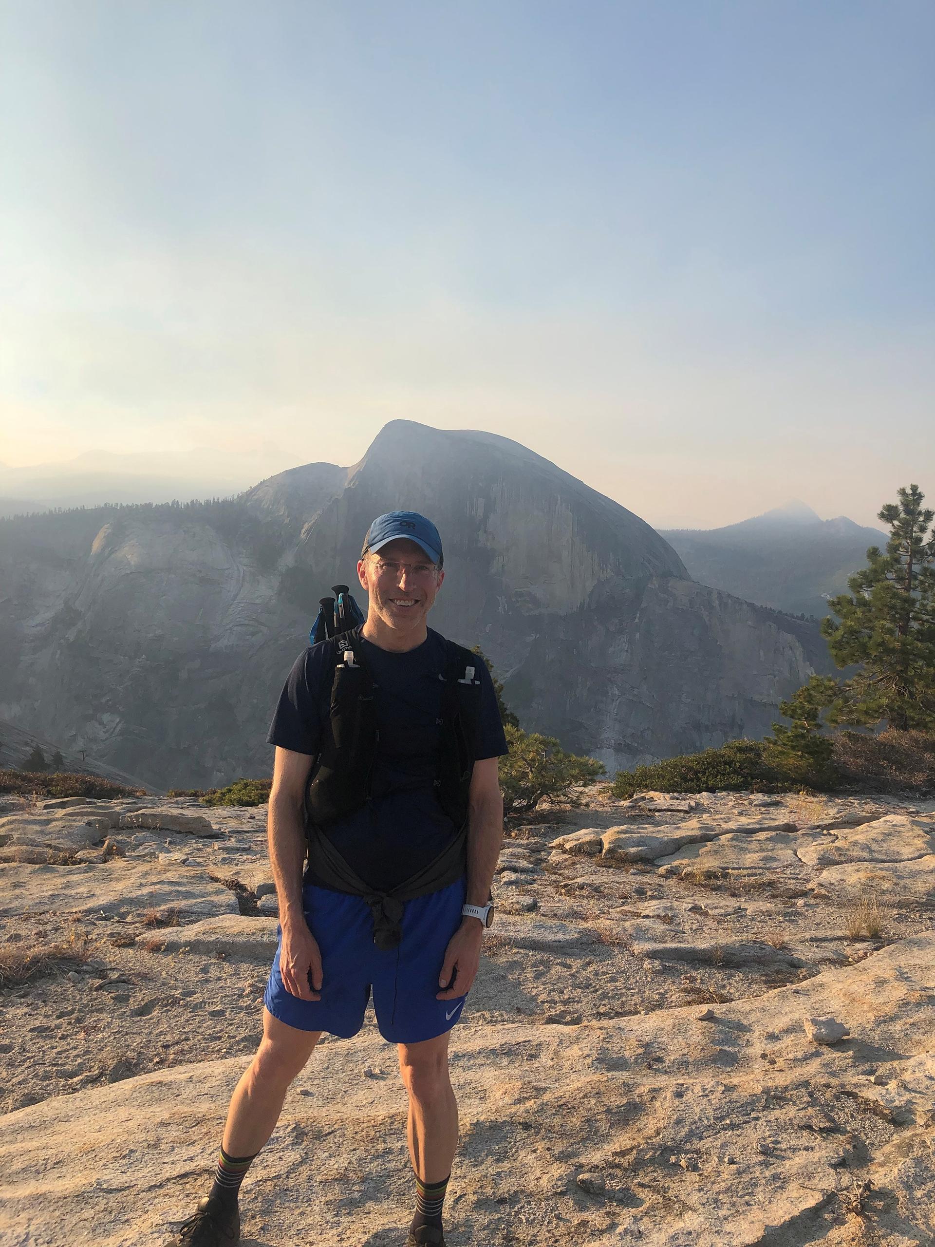 Mark Harrast stands on a lookout point with a mountain rising behind him. 
