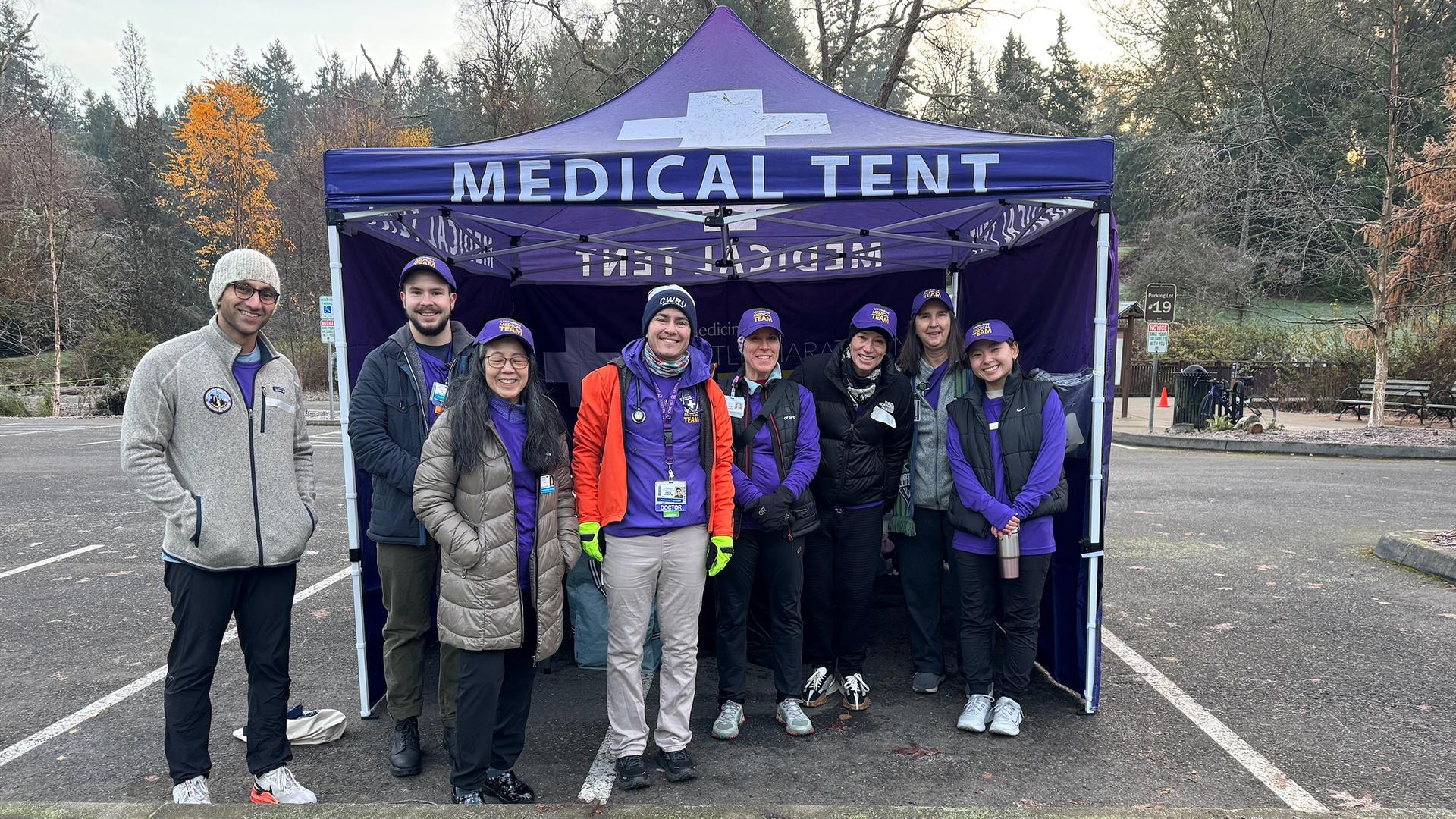 A group of people stand in a parking lot, in front of a purple medical tent. 
