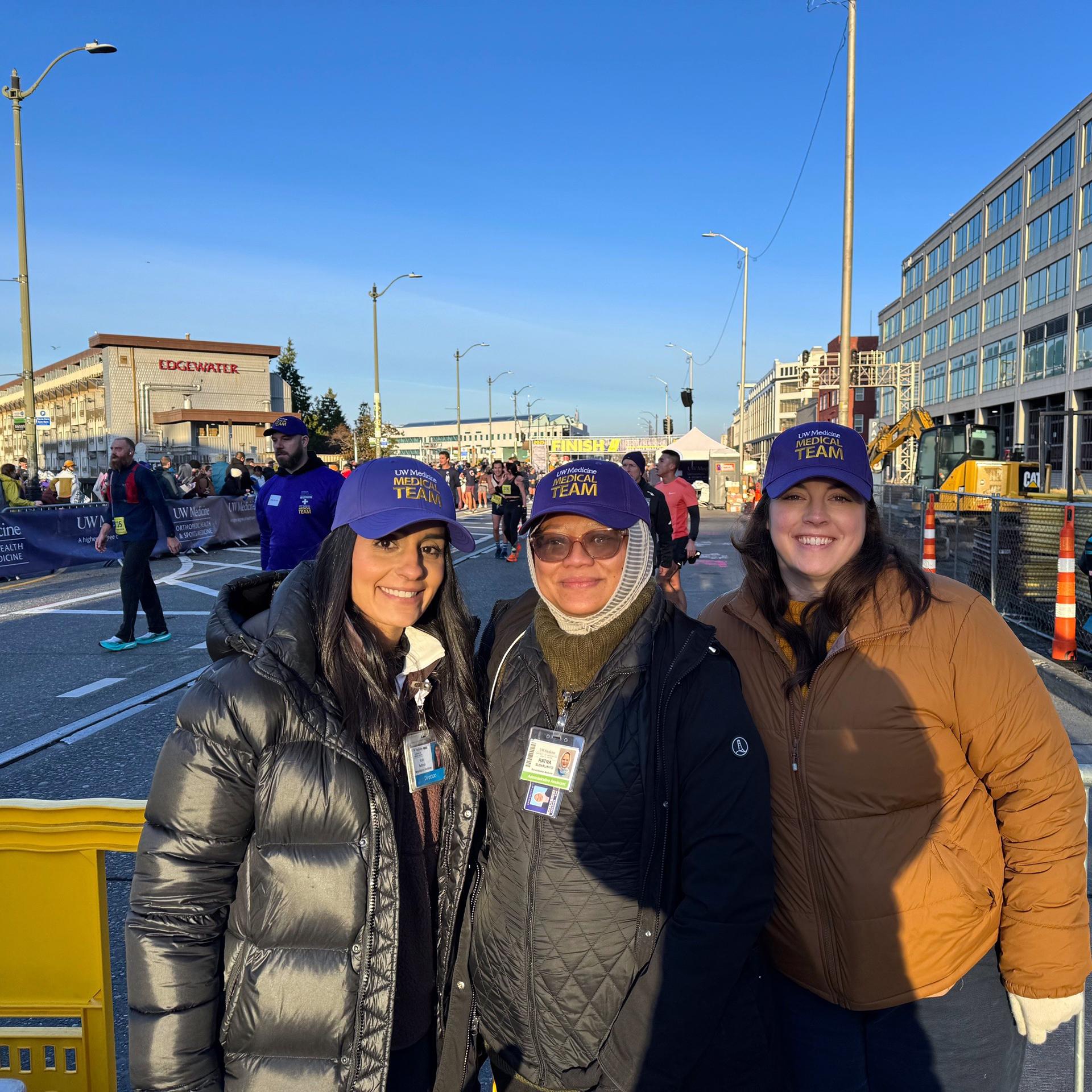 Three women smile for the camera on a sunny day, with a race finish line in the background. 