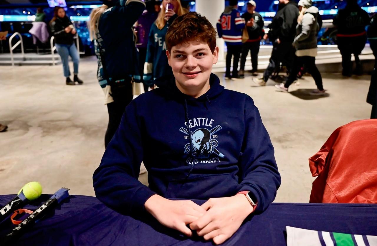 A boy smiles at the camera, wearing a dark blue "Seattle Sled Hockey" t-shirt.