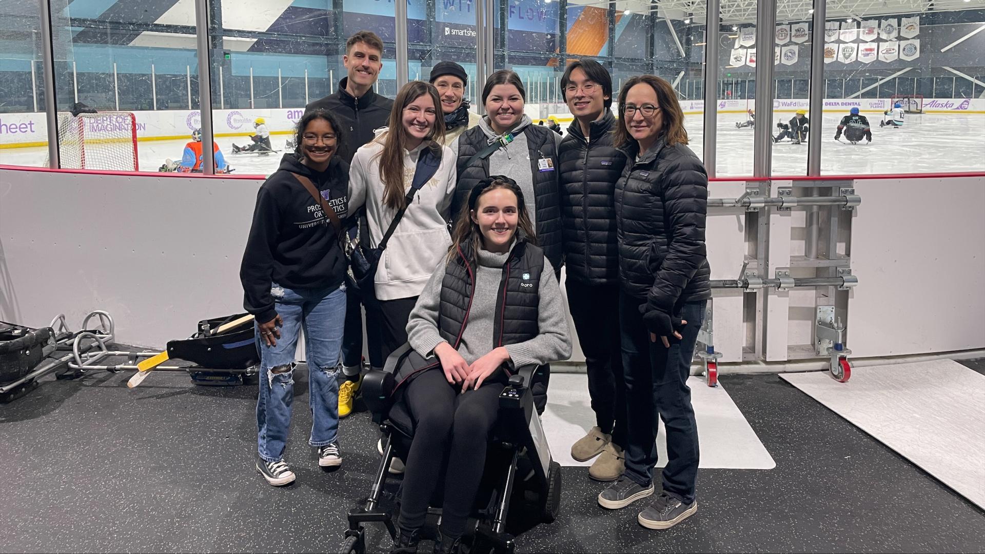 A group of people, one sitting in a sled hockey sled, stand in front of a sled hockey practice at an ice rink. 