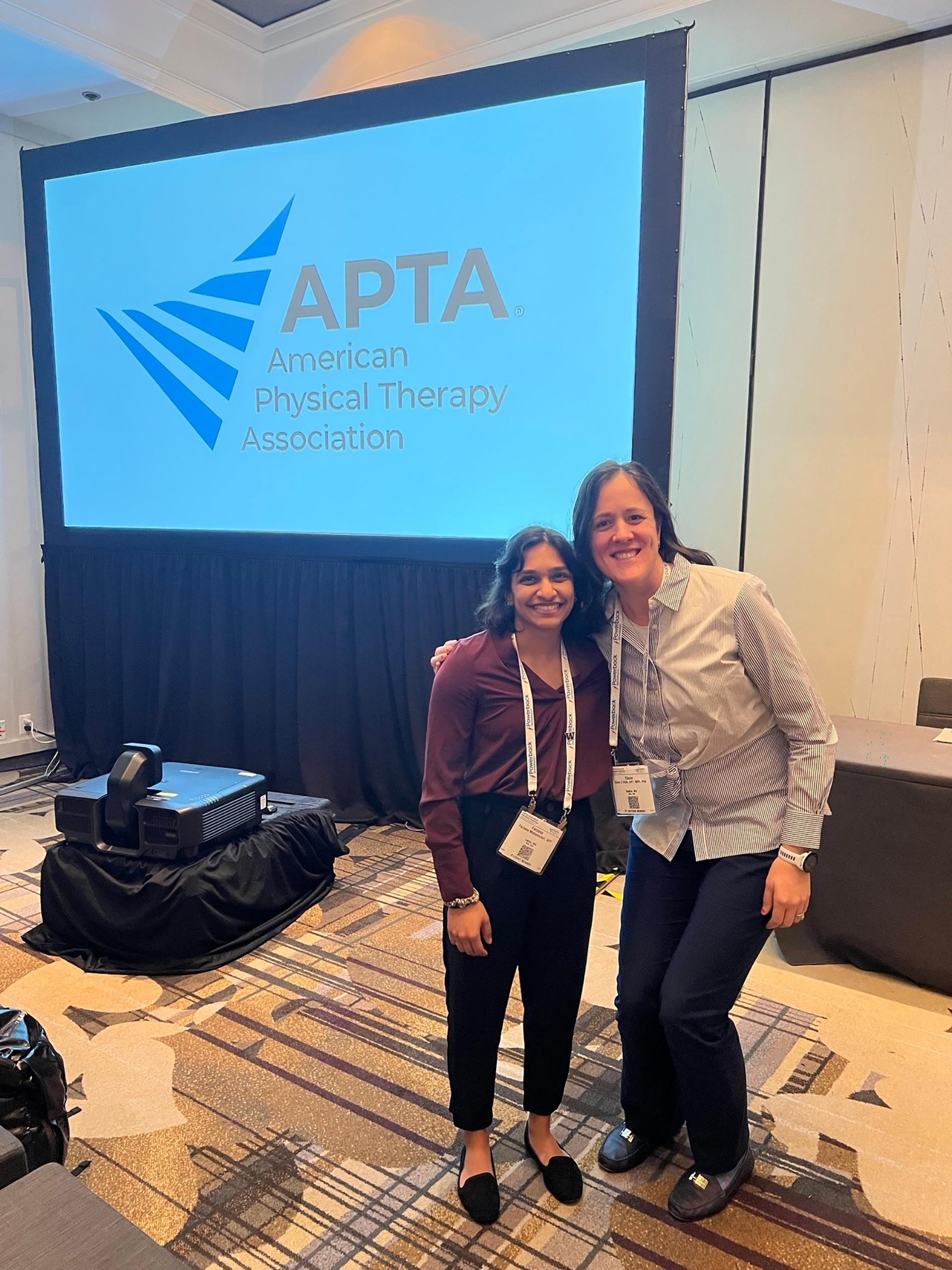 Two people standing together in front of a screen that reads APTA. 
