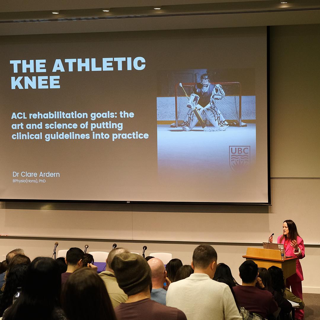 A woman at a podium speaks to a crowd under a slide that reads, "The Athletic Knee."